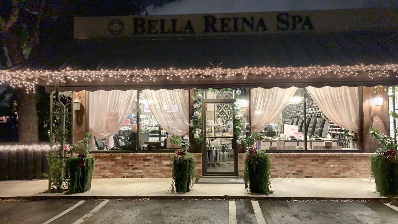 The Bella Reina Spa storefront on George Bush Boulevard at dusk — brick front, cream curtains, planters and string lights