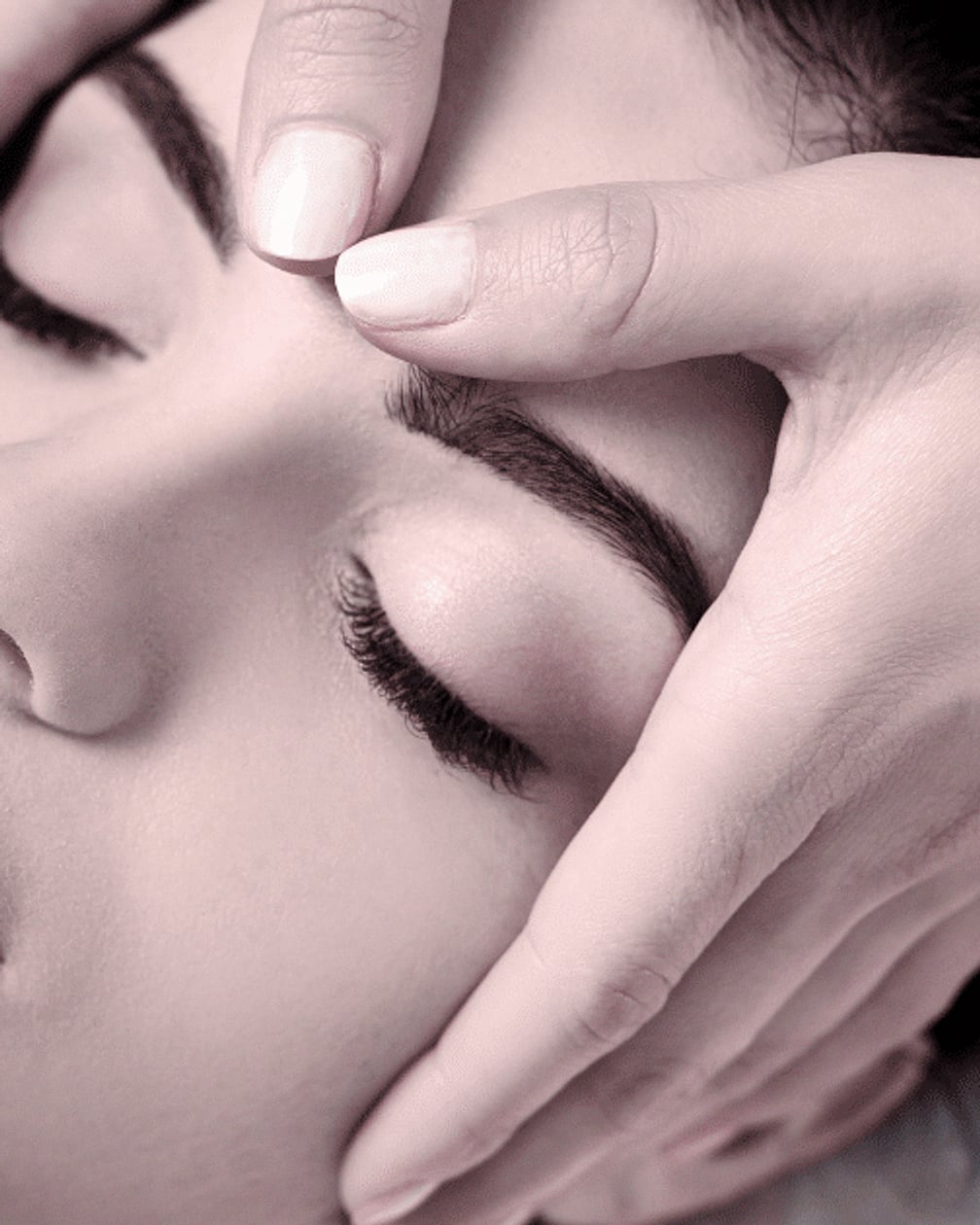 A woman receiving a facial at Bella Reina Spa, hands framing her face in soft pink light
