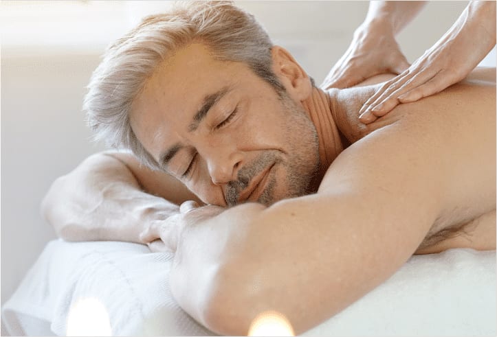 A Bella Reina Spa practitioner applying a body exfoliation product to a client's forearm in a treatment room finished in deep aubergine and porcelain, salt-scrub texture visible on a nearby instrument tray
