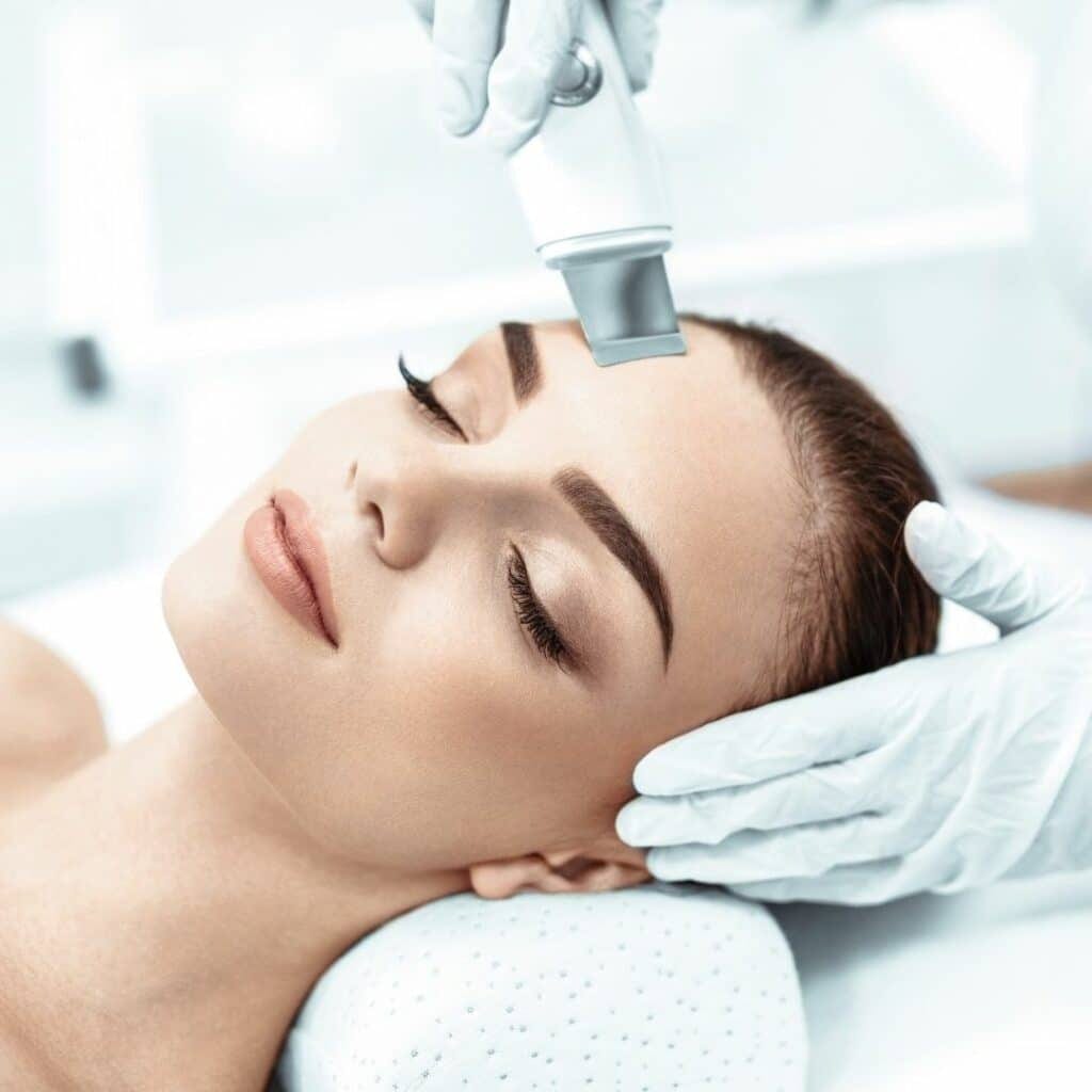 A microdermabrasion wand with a diamond-tip head moving across a client's forehead at Bella Reina Spa, clear tubing carrying exfoliated debris away, treatment room in porcelain and aubergine tones